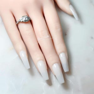 Könnte beinhalten: Ein silberfarbener Ring mit einem quadratischen Mittelstein, der von einem Kranz kleinerer, klarer Steine auf einer dünnen Ringschiene umgeben ist. Der Ring wird an einem Finger mit langen, milchig-weißen Sargnägeln getragen. Der Text Rose Smith ist sichtbar.