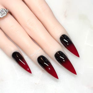 Puede incluir: Un conjunto de uñas artificiales largas y puntiagudas con un diseño degradado de negro a rojo. Las uñas están en una mano de maniquí, con un anillo plateado visible en el dedo índice. Las uñas tienen un acabado brillante.