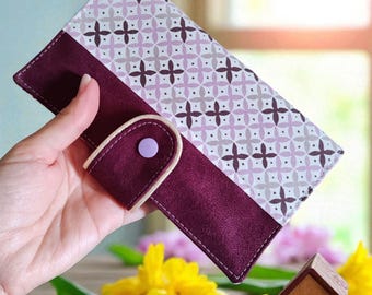 May include: A rectangular wallet featuring a deep plum-colored suede-like lower section and a light lavender and white patterned upper section with a repeating cross motif. The wallet includes a matching plum-colored flap closure with a light purple snap button.