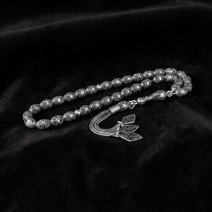 May include: A silver-colored prayer bead rosary with intricately patterned beads. The rosary features a decorative tassel with a geometric design. The beads are approximately 0.5 inches in diameter. The rosary is displayed on a black velvet surface.