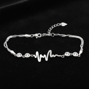 Op de afbeelding: Een zilveren armband met een hartslagontwerp. De armband heeft een fijne ketting met kleine, ronde, heldere edelstenen en een centraal hartslagmotief. De achtergrond is zwart.
