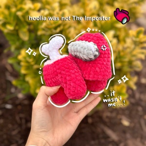 May include: A crocheted red Among Us character with a white bone and a grey visor. The character is being held in a hand with the text "hoolia was not The Impostor" and "...it wasn't me..." written on the image.