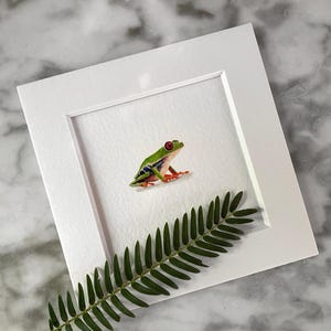 May include: A framed illustration of a green and red tree frog with white and blue markings. The frog is sitting on a white background with a green fern leaf in the foreground.