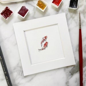 Peut inclure: Un cadre blanc avec un fond blanc et deux poissons rouges et blancs peints à l'aquarelle.