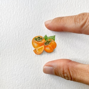 May include: A watercolor painting of three persimmons, two whole and one sliced in half, with green leaves.