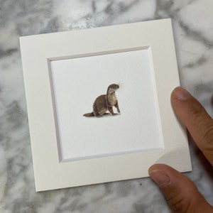 May include: A framed watercolor painting of a brown otter sitting on a white background.