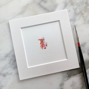 Puede incluir: Una pintura de acuarela de un pez betta con aletas naranja, rojo y blanco. El pez está representado en un marco blanco.