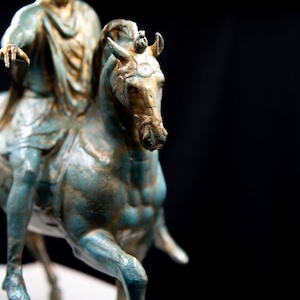 May include: A bronze-toned equestrian statue featuring a figure in classical robes mounted on a horse. The sculpture displays a weathered teal and gold patina finish, with the rider extending one arm forward while seated on the muscular horse.
