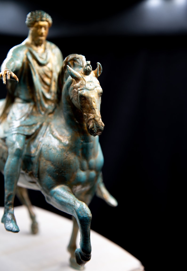 May include: A bronze-toned equestrian statue featuring a figure in classical robes mounted on a horse. The sculpture displays a weathered teal and gold patina finish, with the rider extending one arm forward while seated on the muscular horse.