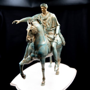 May include: A bronze-toned statue of a figure in classical robes riding a horse. The horse is depicted with one front leg raised, and the rider has one arm extended forward. The sculpture features a weathered, verdigris-like finish with gold highlights.