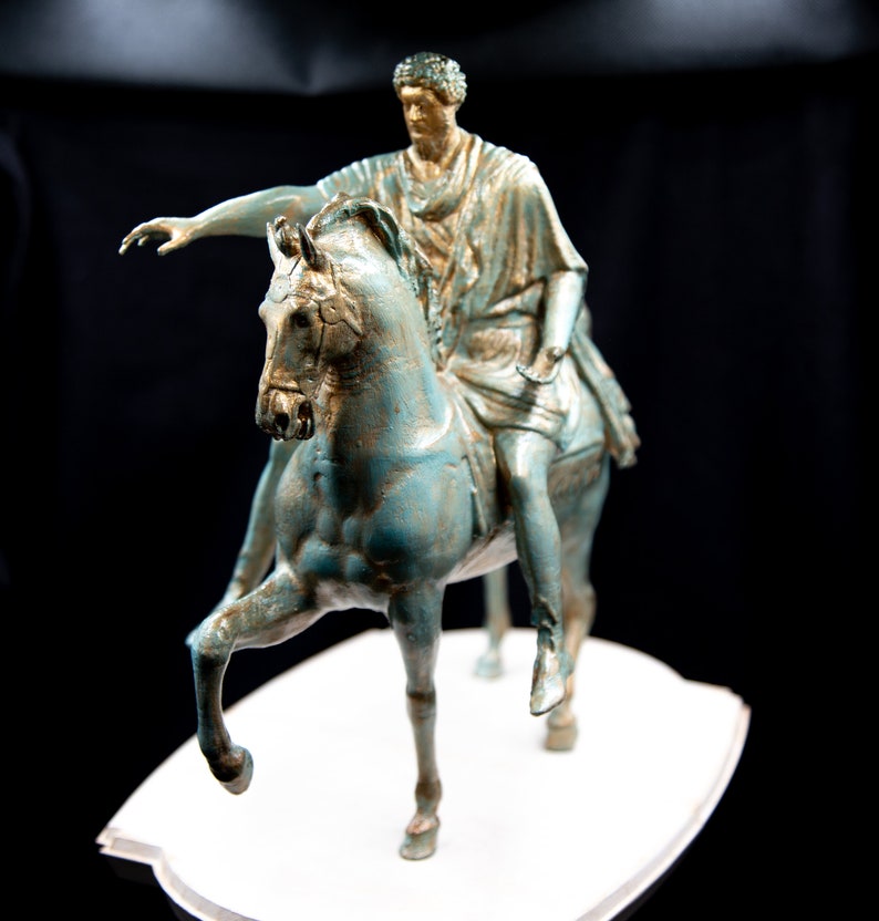 May include: A bronze-toned statue of a figure in classical robes riding a horse. The horse is depicted with one front leg raised, and the rider has one arm extended forward. The sculpture features a weathered, verdigris-like finish with gold highlights.