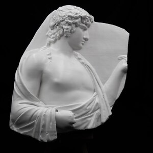 May include: A white plaster sculpture of a classical figure, bust-length, against a black background. The figure has curly hair and is draped in a cloth. The sculpture is a relief, with the figure emerging from a flat plane.
