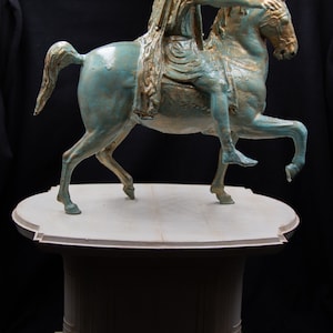 May include: A bronze-toned equestrian statue featuring a figure in a draped robe riding a horse with one leg raised. The sculpture has a weathered teal and gold finish and is mounted on a light-coloured, tiered, circular pedestal base.