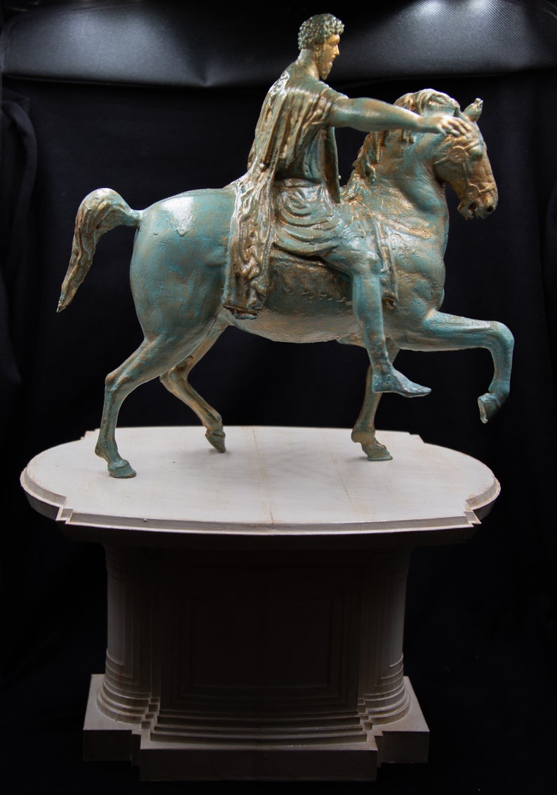 May include: A bronze-toned equestrian statue featuring a figure in a draped robe riding a horse with one leg raised. The sculpture has a weathered teal and gold finish and is mounted on a light-coloured, tiered, circular pedestal base.