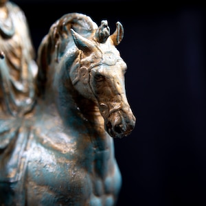 May include: A weathered bronze-toned statue of a horse and rider. The horse features a textured finish with teal and gold highlights, a bridle, and a mane. The rider is partially visible, wearing draped clothing and extending one arm forward.