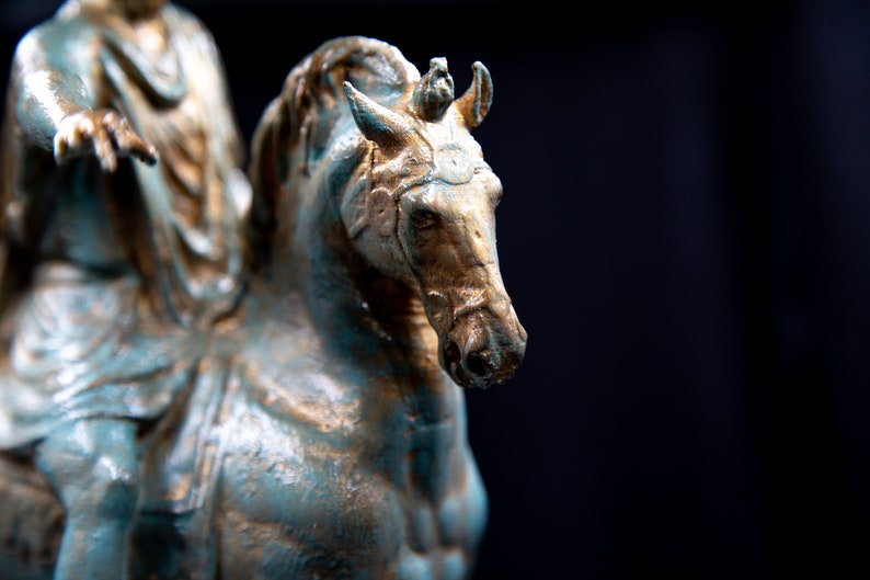 May include: A weathered bronze-toned statue of a horse and rider. The horse features a textured finish with teal and gold highlights, a bridle, and a mane. The rider is partially visible, wearing draped clothing and extending one arm forward.