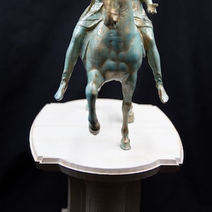 May include: A bronze-toned statue of a figure riding a horse, featuring a weathered teal and gold finish. The rider is depicted with one arm extended forward, mounted on a horse in a trotting pose. The entire sculpture is set upon a light-coloured, tiered pedestal base.