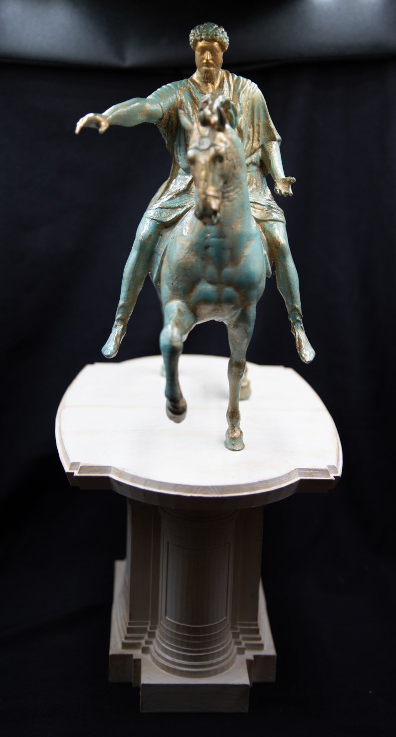 May include: A bronze-toned statue of a figure riding a horse, featuring a weathered teal and gold finish. The rider is depicted with one arm extended forward, mounted on a horse in a trotting pose. The entire sculpture is set upon a light-coloured, tiered pedestal base.