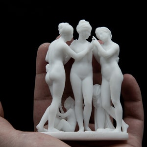 May include: White ceramic sculpture of three figures standing together. The figures are nude and appear to be women. The sculpture is detailed and has a smooth finish.