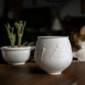 Può includere: Un vaso di ceramica bianca con un design in rilievo di figure e volute. Il vaso è appoggiato su un tavolo di legno.