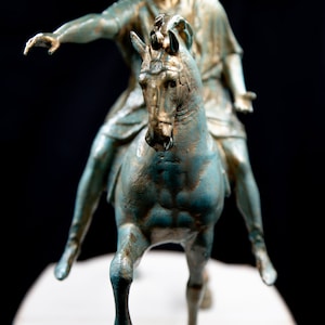 May include: A bronze-toned statue of a figure riding a horse, featuring a weathered teal and gold finish. The rider is depicted with one arm extended forward and the other held out to the side, while the horse is shown in a forward-moving pose.