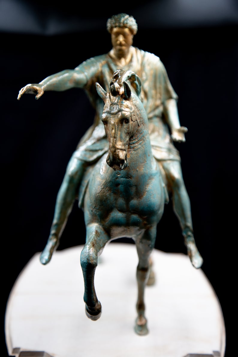 May include: A bronze-toned statue of a figure riding a horse, featuring a weathered teal and gold finish. The rider is depicted with one arm extended forward and the other held out to the side, while the horse is shown in a forward-moving pose.