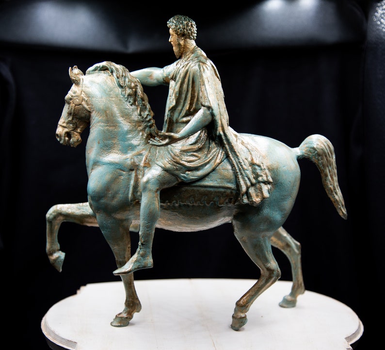 May include: A teal and gold-toned equestrian statue depicting a figure in a draped robe riding a horse with one front leg raised. The sculpture features a weathered finish with metallic gold highlights on the horse's mane and the rider's clothing.