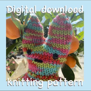May include: A colorful knitted bunny with black button eyes and a stitched mouth. The bunny is set against a backdrop of orange fruit and green leaves. Text at the top reads "Digital download" and at the bottom "knitting pattern".