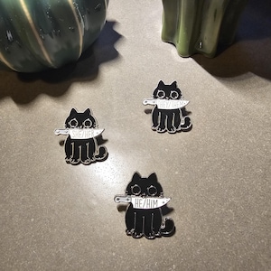 May include: Three black cat enamel pins with a knife in their mouths. Each pin has a different pronoun on the knife: "She/Her", "He/Him", and "They/Them".