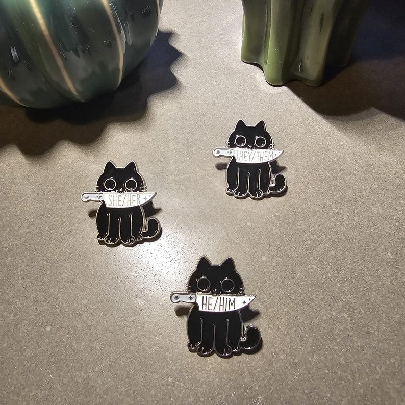 May include: Three black cat enamel pins with a knife in their mouths. Each pin has a different pronoun on the knife: "She/Her", "He/Him", and "They/Them".