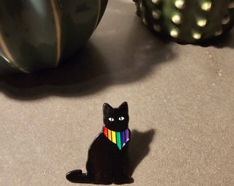 May include: Black enamel cat pin with a rainbow colored scarf. The cat is sitting with its head tilted slightly to the side.