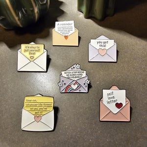 Puede incluir: Una colección de pines de esmalte con forma de sobres, cada uno con un mensaje único. Los pines presentan varios colores y diseños, incluyendo corazones, arcoíris y detalles florales. Los mensajes incluyen "It's okay to put yourself first" y "You got this!"