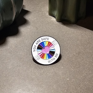 Puede incluir: Un pin redondo de esmalte con el texto "YOU ARE SAFE WITH ME". El pin presenta un diseño de arcoíris con un corazón rosa en el centro, rodeado de manos blancas. El pin está sobre una superficie gris.