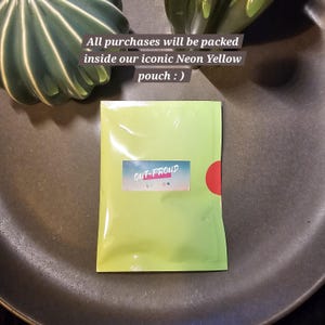May include: A bright green pouch with the text "OUT-PROUD LONDON" printed on it. The pouch is sealed and has a red circle on the right side. The text "All purchases will be packed inside our iconic Neon Yellow pouch :)" is written above the pouch.