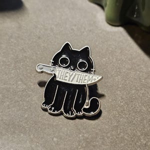 May include: Black enamel pin of a cat holding a knife in its mouth. The knife has the words "They/Them" written on it.