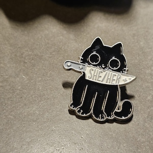 May include: Black enamel pin of a cat with a knife in its mouth. The knife has the words "SHE/HER" written on it.