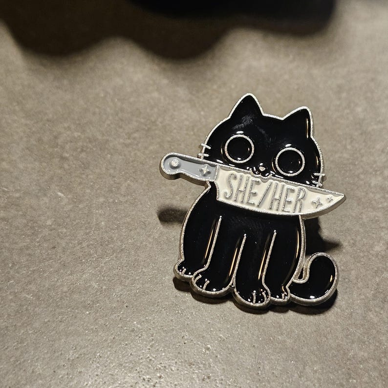May include: Black enamel pin of a cat with a knife in its mouth. The knife has the words "SHE/HER" written on it.