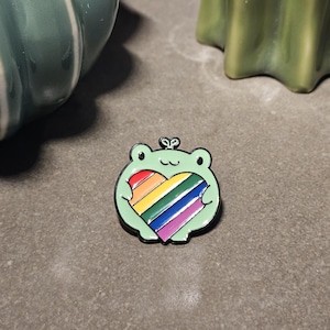 Puede incluir: Un pin de esmalte verde claro con forma de rana que sostiene un corazón con rayas diagonales en rojo, naranja, amarillo, verde, azul y morado. La rana tiene contornos negros, dos ojos pequeños y un pequeño brote en la parte superior.