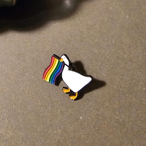 Peut inclure: Une broche en émail blanc en forme d'oie avec un bec et des pattes orange. L'oie tient dans son bec un petit drapeau rectangulaire aux couleurs de l'arc-en-ciel. La broche présente des contours noirs et une finition brillante.