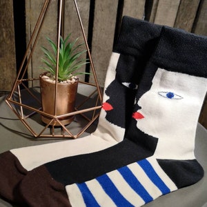 May include: A pair of black and white socks with a graphic print of two faces. The faces are black and white with red lips and blue eyes. The socks have a blue and white striped pattern at the bottom.