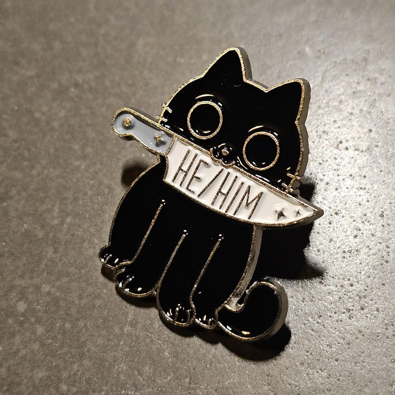 May include: Black enamel pin of a cartoon cat holding a knife with the text "HE/HIM" on the blade.