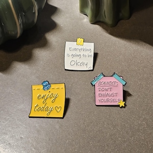 Op de afbeelding: Drie emaille pins met motiverende berichten. Eén is een gele notitie met de tekst "enjoy today" met een hartje. Een andere is een witte notitie met de tekst "Everything is going to be Okay". De derde is een roze huisvormige pin met de tekst "Reminder: Don't Exhaust Yourself".