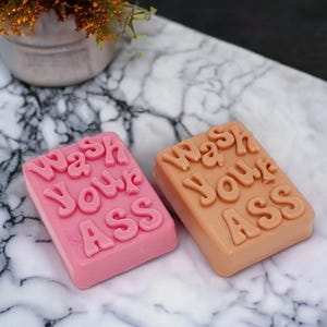 May include: Two rectangular bars of soap, one pink and one tan, each featuring the embossed text "Wash Your Ass" in a bold, rounded font. The soap bars are solid in colour with a smooth finish, displaying clear, raised lettering on the top surface.