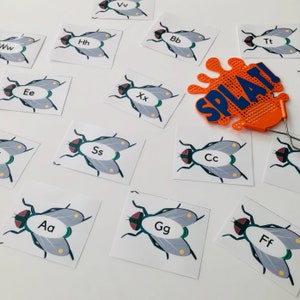May include: A set of 12 fly swatter game cards featuring different letters of the alphabet. Each card has a cartoon fly with the letter printed on it. The cards are printed on white cardstock and have a blue border. The cards are arranged in a random order on a white surface. There is also an orange fly swatter with the word "SPLAT!" printed on it.