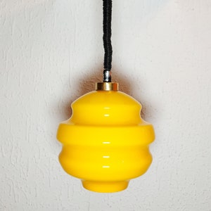 Könnte beinhalten: Eine gelbe Glaspendelleuchte mit schwarzem Kabel und schwarzer Deckenbefestigung.