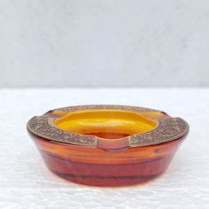 Może przedstawiać: Mały popielniczkę ze szkła bursztynowego z ozdobnym wzorem w kolorze złotym wokół krawędzi.