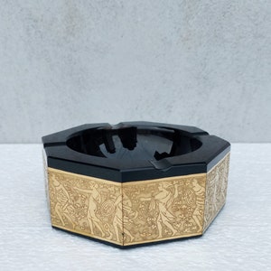 Może przedstawiać: Czarny, ośmiokątny popielniczkę z pozłacanym rantem i ozdobnym wzorem przedstawiającym figury w reliefie. Wzór przypomina starożytną mitologię grecką.