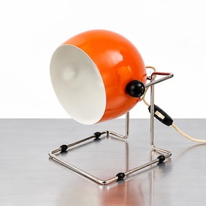 Könnte beinhalten: Eine leuchtend orangefarbene, kugelförmige Tischleuchte mit weißem Innenschirm. Die Lampe verfügt über einen Rahmen aus poliertem Chrommetall mit schwarzen, kugelförmigen Akzenten an den Gelenken und einen schwarzen Schalter am hellen Kabel.