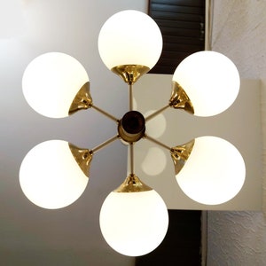 K&ouml;nnte beinhalten: Ein sechsarmiger Kronleuchter mit wei&szlig;en Glaskugeln und goldenen Metallakzenten. Der Kronleuchter hat ein sternenf&ouml;rmiges Design, wobei die Kugeln in einem Kreis um einen zentralen Punkt angeordnet sind.