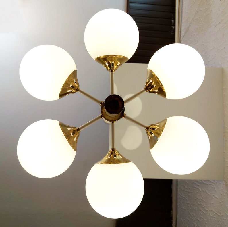 K&ouml;nnte beinhalten: Ein sechsarmiger Kronleuchter mit wei&szlig;en Glaskugeln und goldenen Metallakzenten. Der Kronleuchter hat ein sternenf&ouml;rmiges Design, wobei die Kugeln in einem Kreis um einen zentralen Punkt angeordnet sind.